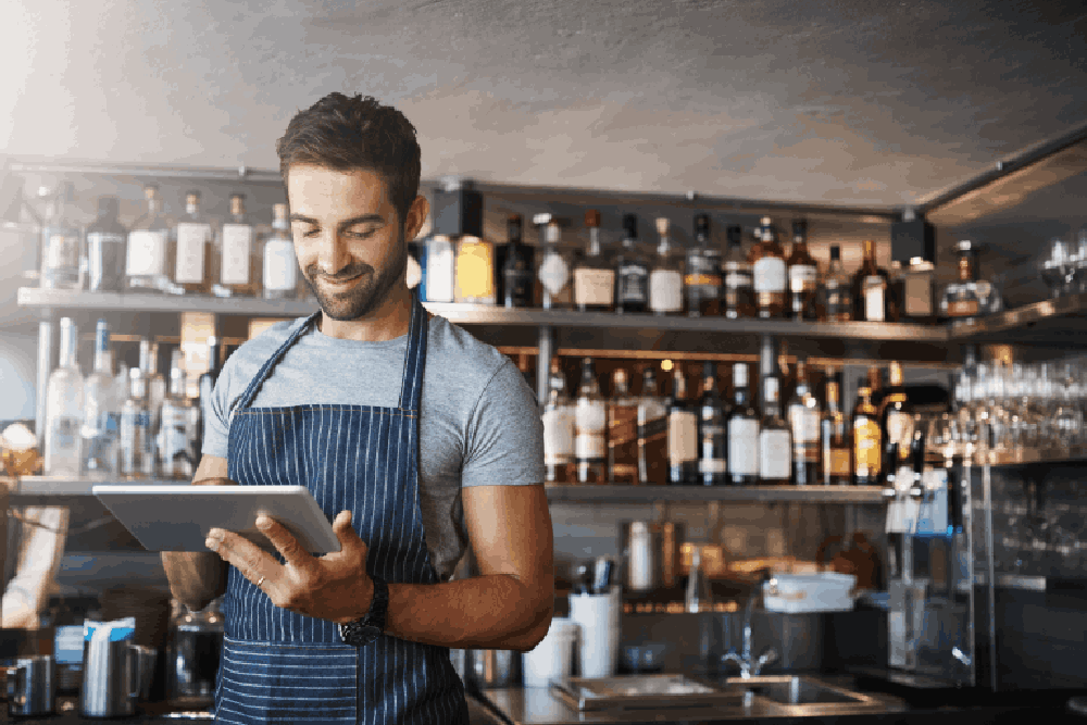 Person in blue apron holding a tablet in a bar or restaurant setting with various bottles and glasses on the shelves.