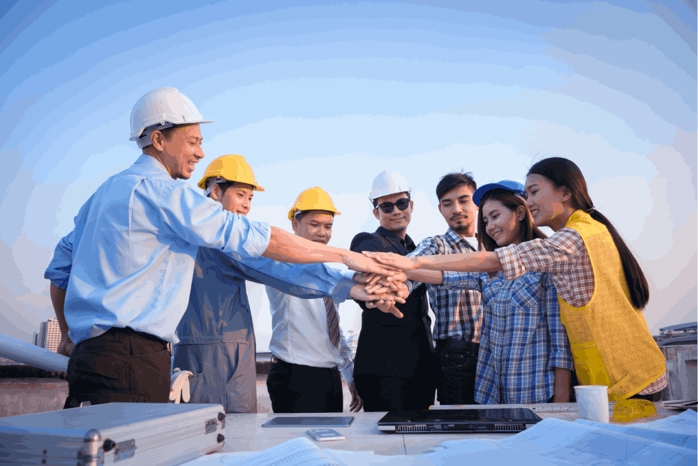 Team of construction workers in hard hats and safety vests on a rooftop, standing in a circle with a laptop and papers on the table, showing team trustness.