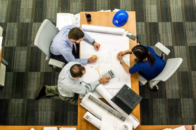 Three professionals analyzing a blueprint on a table in an office, with a hard hat and coffee cup nearby.